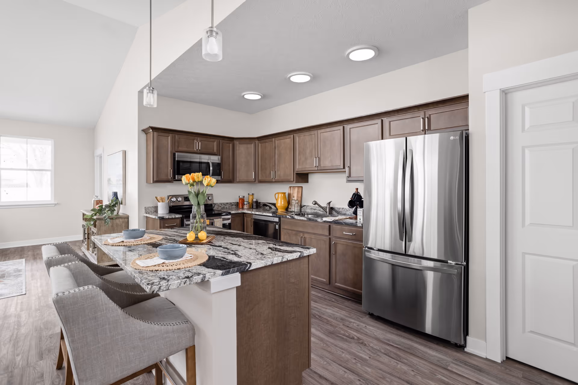 Modern kitchen with a marble-topped island, pendant lights, gray upholstered bar stools, stainless steel refrigerator, and wooden cabinets.