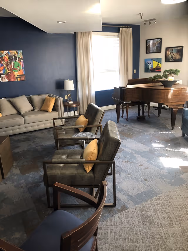 A cozy senior living common area with a gray sofa, three armchairs with yellow cushions, a wooden coffee table, and a grand piano. The room has blue walls, a window with white curtains, and colorful paintings on the walls.