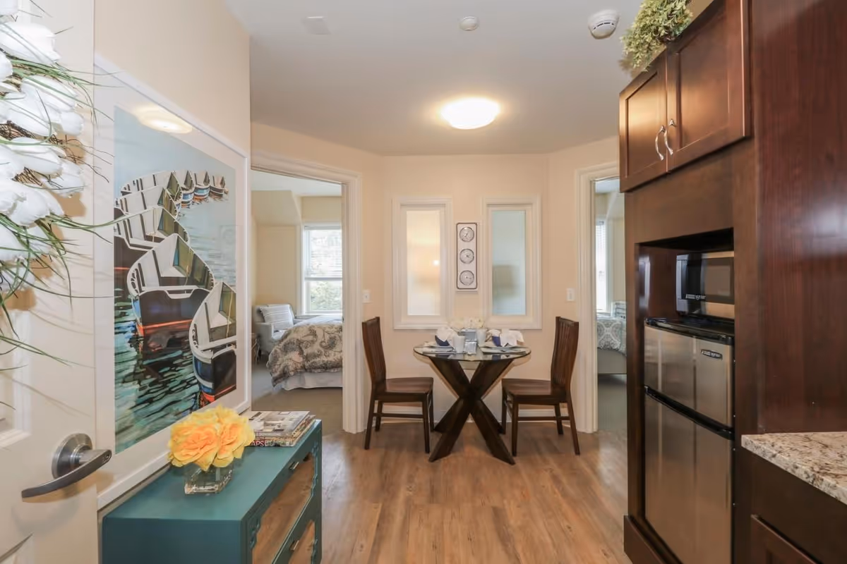 Small dining nook with a round glass-top table and two wooden chairs adjacent to a kitchenette and bedroom doorways.