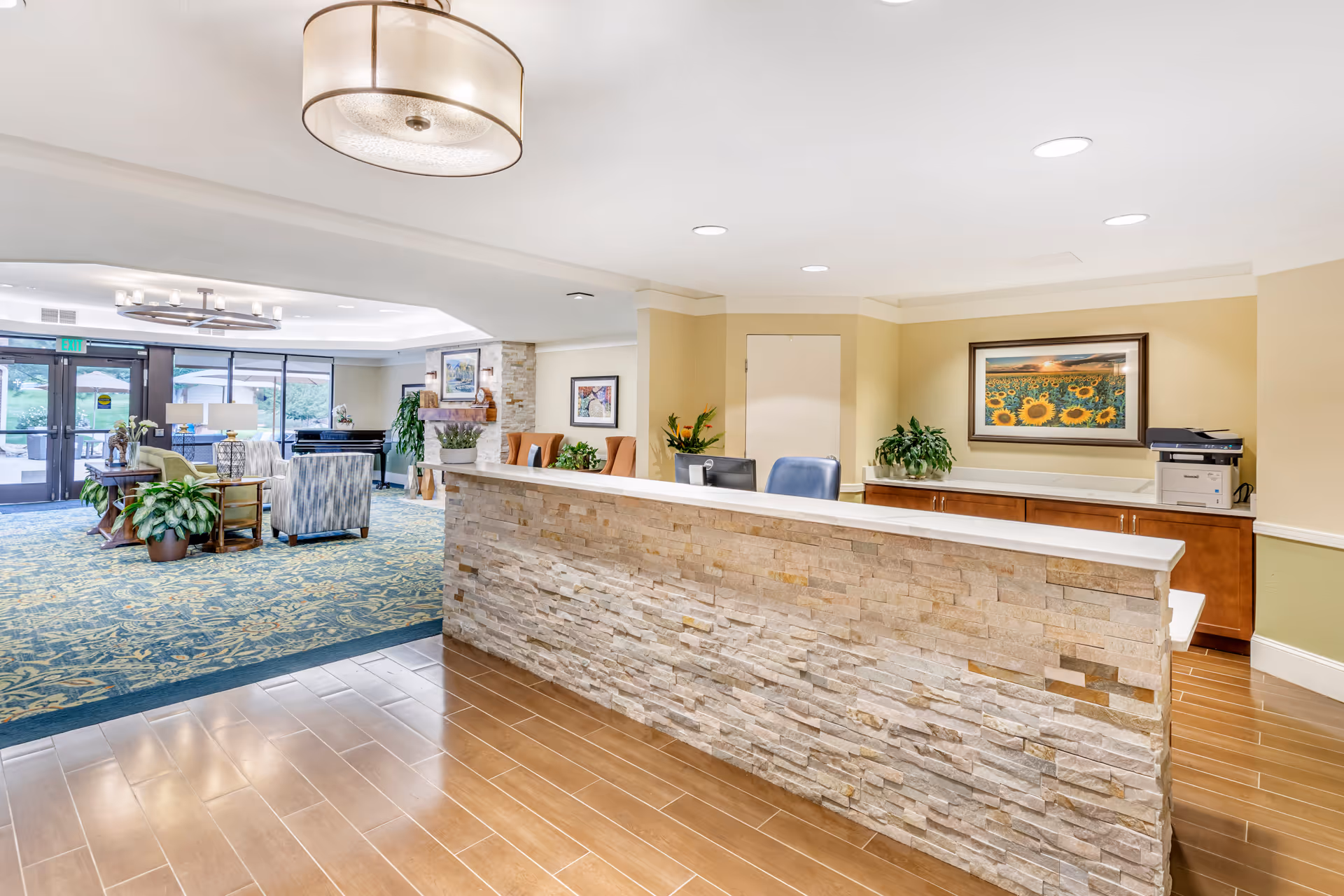 Reception area of Brookdale Meridian Boulder featuring a stone-faced front desk with two chairs behind it, a printer on a wooden cabinet, framed artwork of sunflowers on the wall, and a seating area with chairs and plants near large glass doors leading outside.