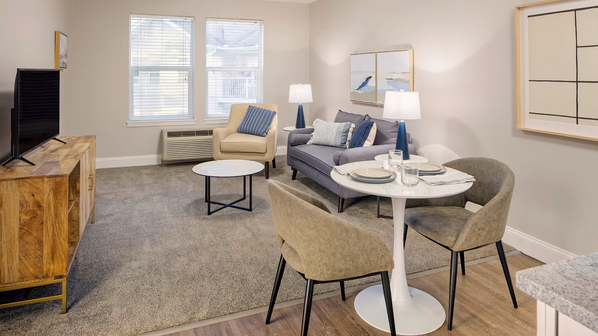 A cozy living room area with a gray sofa, beige armchair, and a round white coffee table on a gray carpet. There is a small dining table set with two chairs, plates, and glasses. The room has two windows with blinds, a wooden TV stand with a flat-screen TV, and two table lamps on either side of the sofa. The walls are decorated with modern artwork.