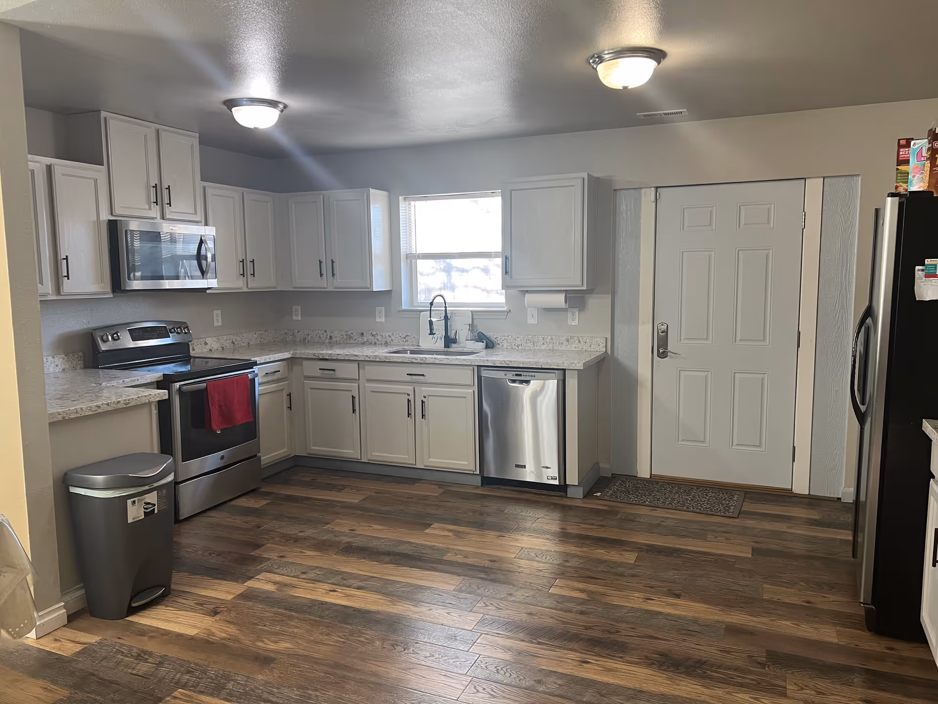 A modern kitchen with white cabinets, stainless steel appliances including a stove, microwave, dishwasher, and refrigerator. There is a window above the sink, wood-style flooring, and a white door with a small mat in front of it. The kitchen is well-lit with ceiling lights.