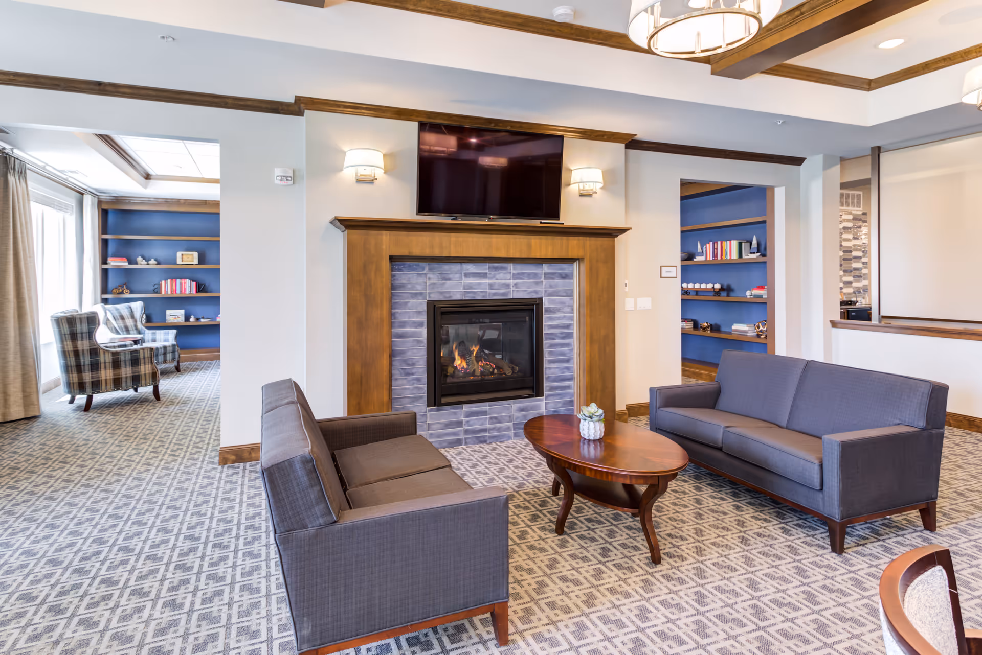 A cozy senior living common area with two gray sofas facing a wooden coffee table and a fireplace with blue tiles. Above the fireplace is a mounted flat-screen TV. The room has patterned carpet, wall sconces, and wooden trim. In the background, there are built-in blue shelves with books and decorative items, and two plaid armchairs near a window with curtains.