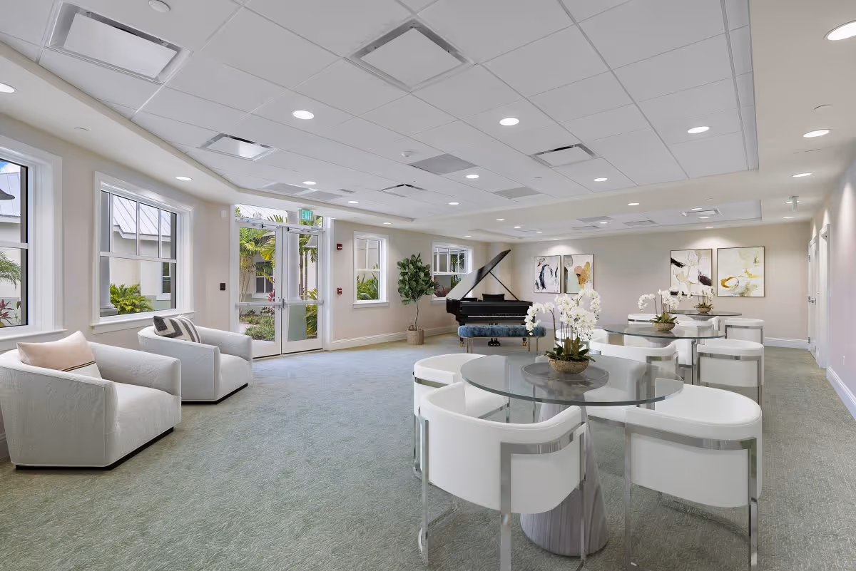 Bright senior living common area with round glass dining tables and white chairs, a grand piano, and lounge seating near windows.