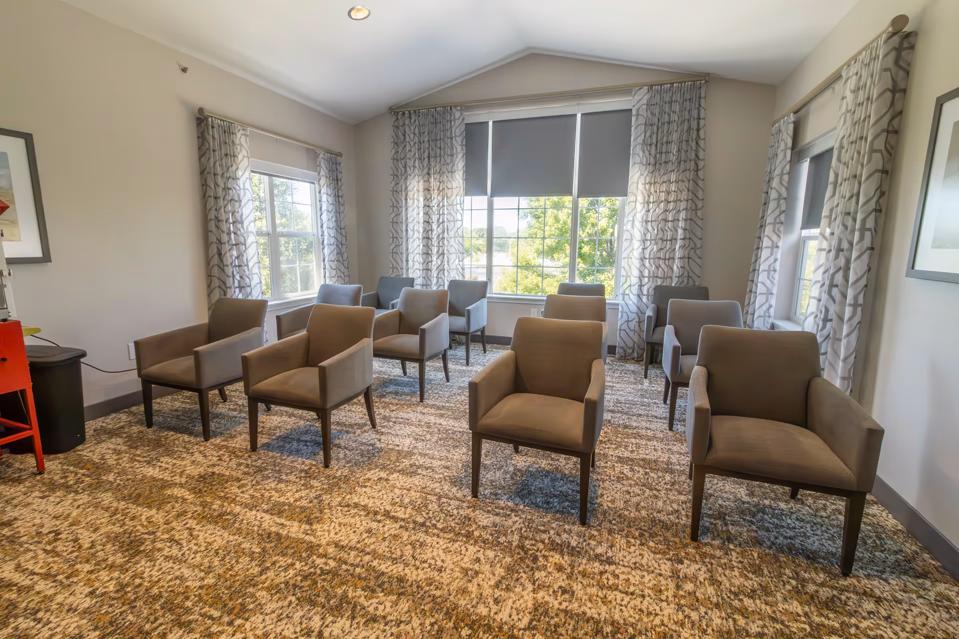 A bright room with multiple gray armchairs arranged in rows facing forward. The room has large windows with patterned curtains allowing natural light to enter. The walls are light-colored, and the floor is covered with a textured carpet. There is a red chair and a small black trash bin in the corner.