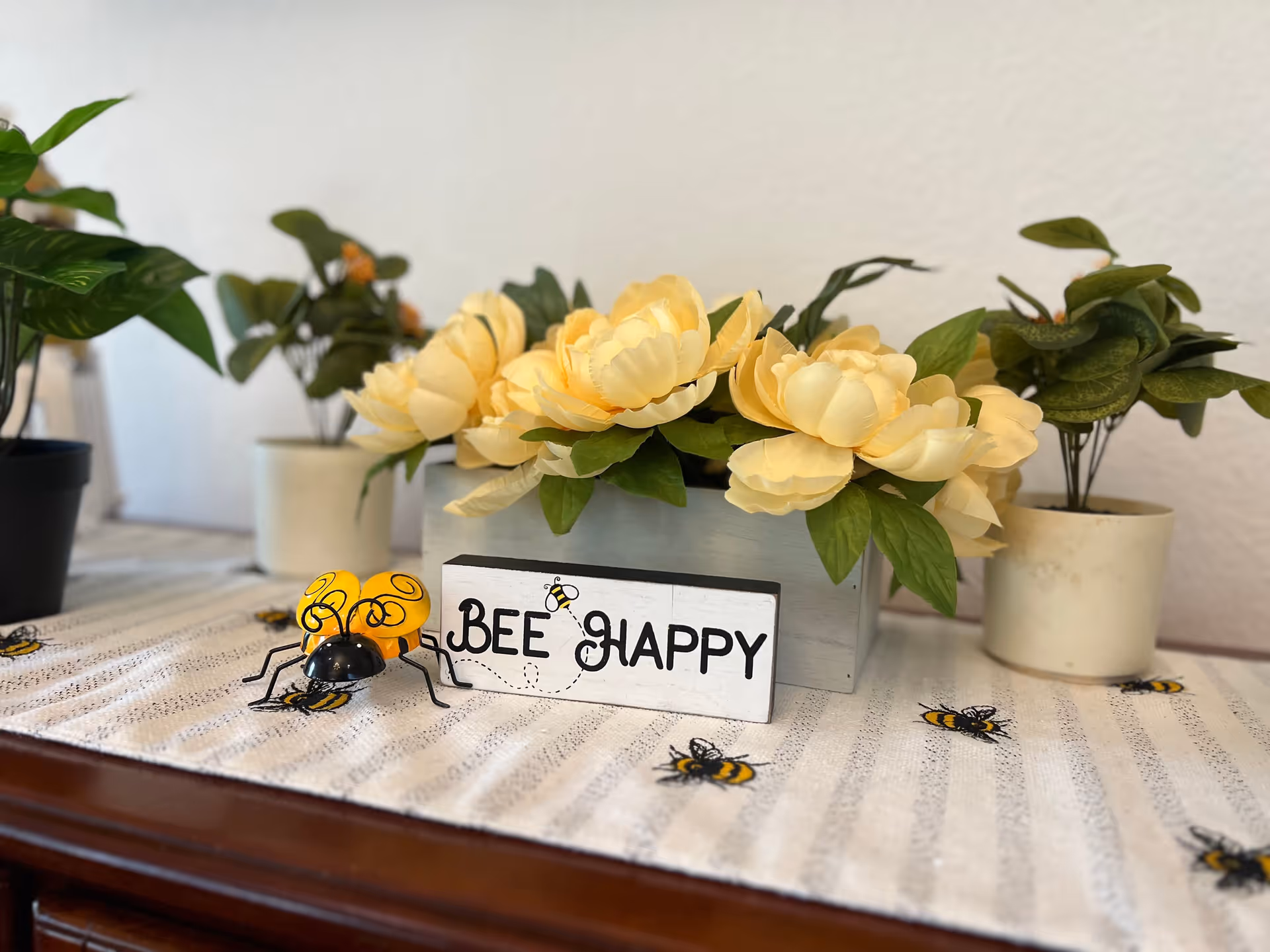 A decorative arrangement on a wooden surface featuring a small sign that reads 'BEE HAPPY' with a bee illustration, surrounded by artificial yellow flowers in a gray rectangular pot and green leafy plants in white pots. The surface is covered with a white cloth decorated with small bee designs.