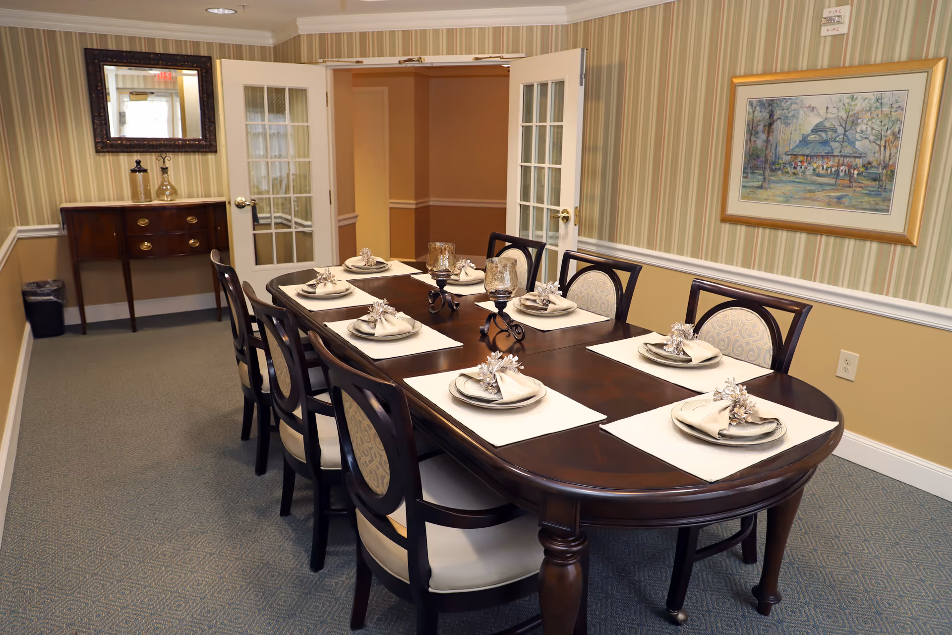 A formal dining room with a long dark wooden table set for six people. Each place setting includes a plate, a beige napkin with a decorative napkin ring, and a placemat. Two decorative candle holders are placed in the center of the table. The room has striped wallpaper with a chair rail, a framed painting on the wall, a mirror above a wooden side table, and double glass-paneled doors leading to another room.