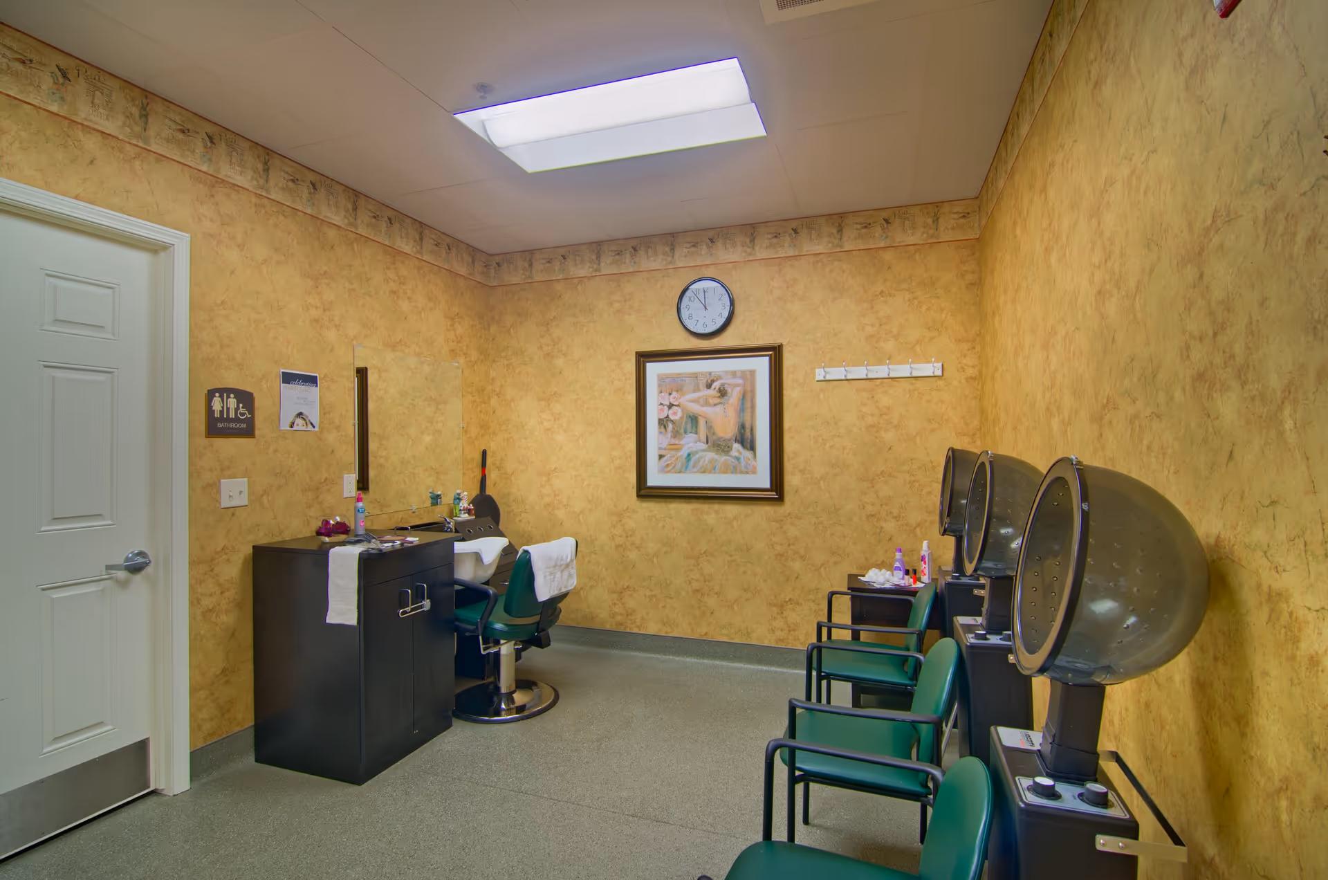 Interior view of a salon or beauty room with yellow textured walls, a white door on the left, a black styling station with a mirror and a green salon chair. On the right side, there are three green chairs with hair dryers attached. A clock and a framed painting hang on the wall.