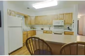 A kitchen with light wood cabinets, a white refrigerator, white microwave, and white stove. There is a countertop with two wooden chairs in the foreground and a hardwood floor.
