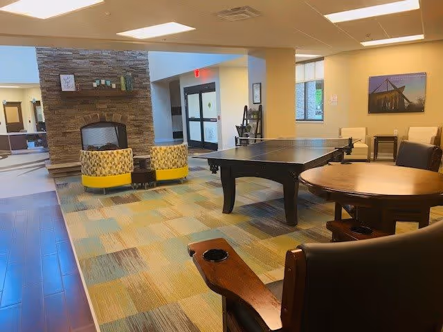 A spacious common area in a senior living facility featuring a stone fireplace, two patterned armchairs with yellow bases, a ping pong table, and several wooden tables and chairs. The room has a colorful patterned carpet and large windows allowing natural light.