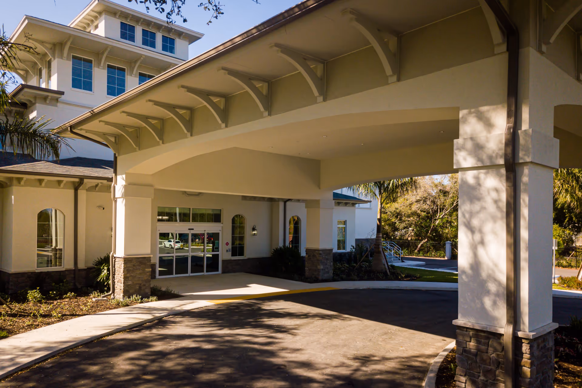 Entrance area of Aravilla Clearwater facility showing a covered driveway with large white pillars and stone bases, glass double doors, and windows. Palm trees and landscaping are visible around the building under a clear sky.