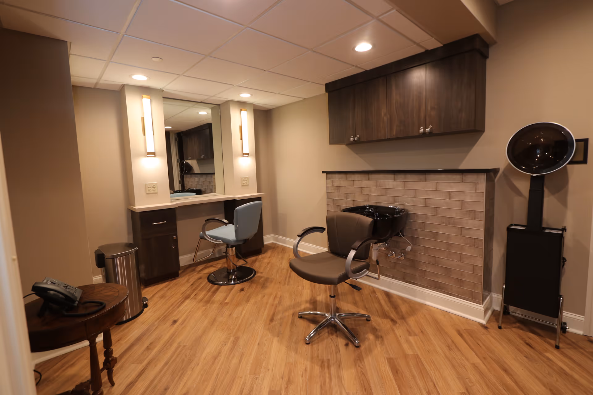 Interior salon room with styling chairs, a mirror and vanity, a hair-washing sink, hooded dryer, and cabinets.
