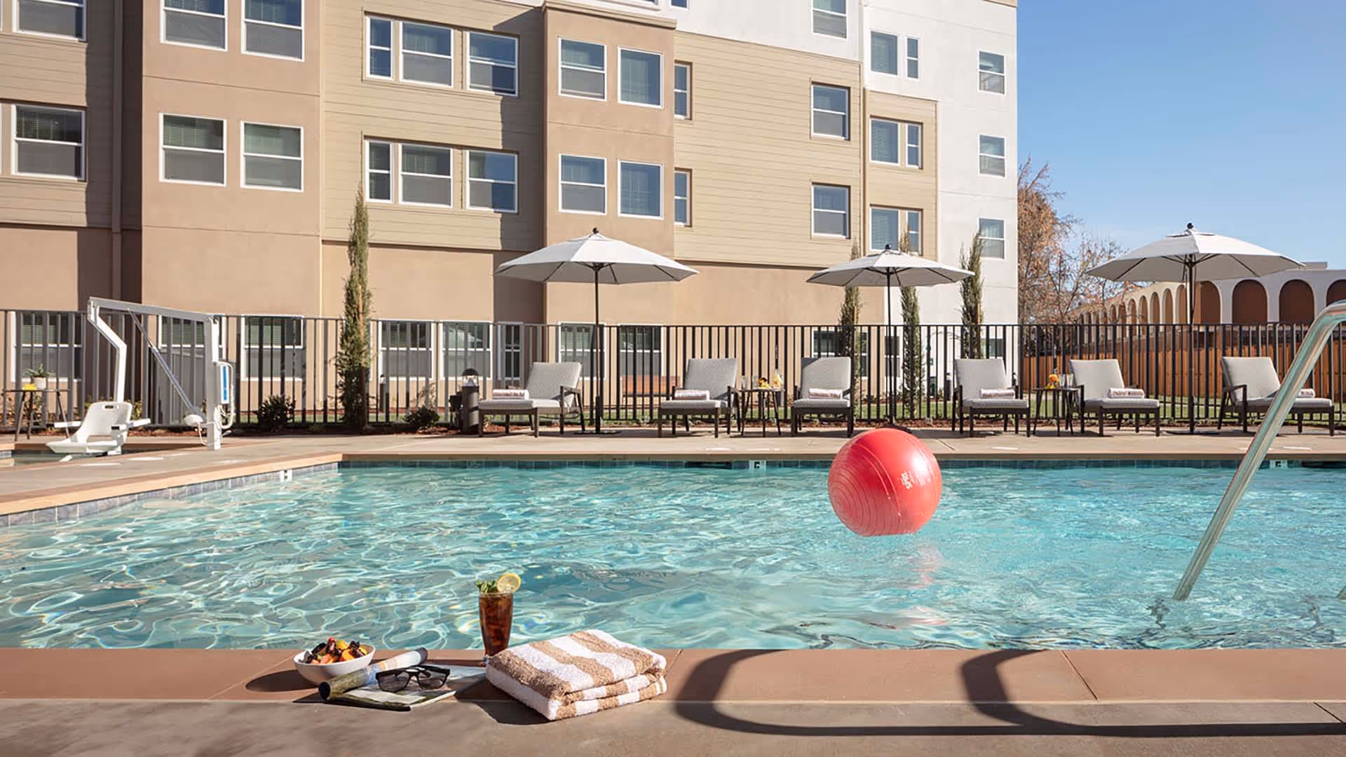 Outdoor swimming pool at a senior living facility with lounge chairs and umbrellas, a red beach ball in the water, and a towel and drink on the pool edge.
