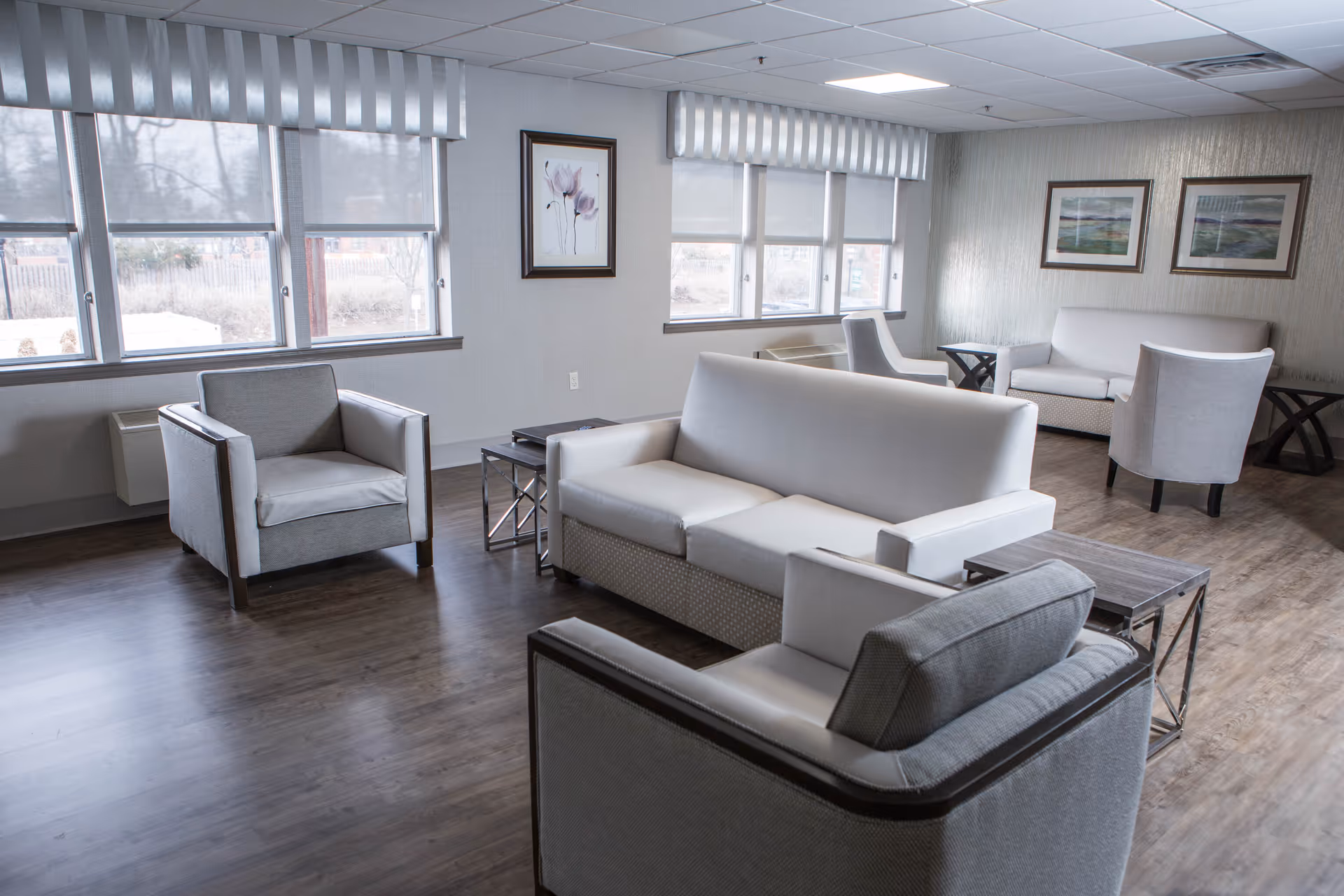 A bright and spacious living room area with large windows letting in natural light. The room features modern furniture including two armchairs, a sofa, and a loveseat arranged around two small coffee tables. The walls are decorated with framed artwork, and the floor has a wood finish.