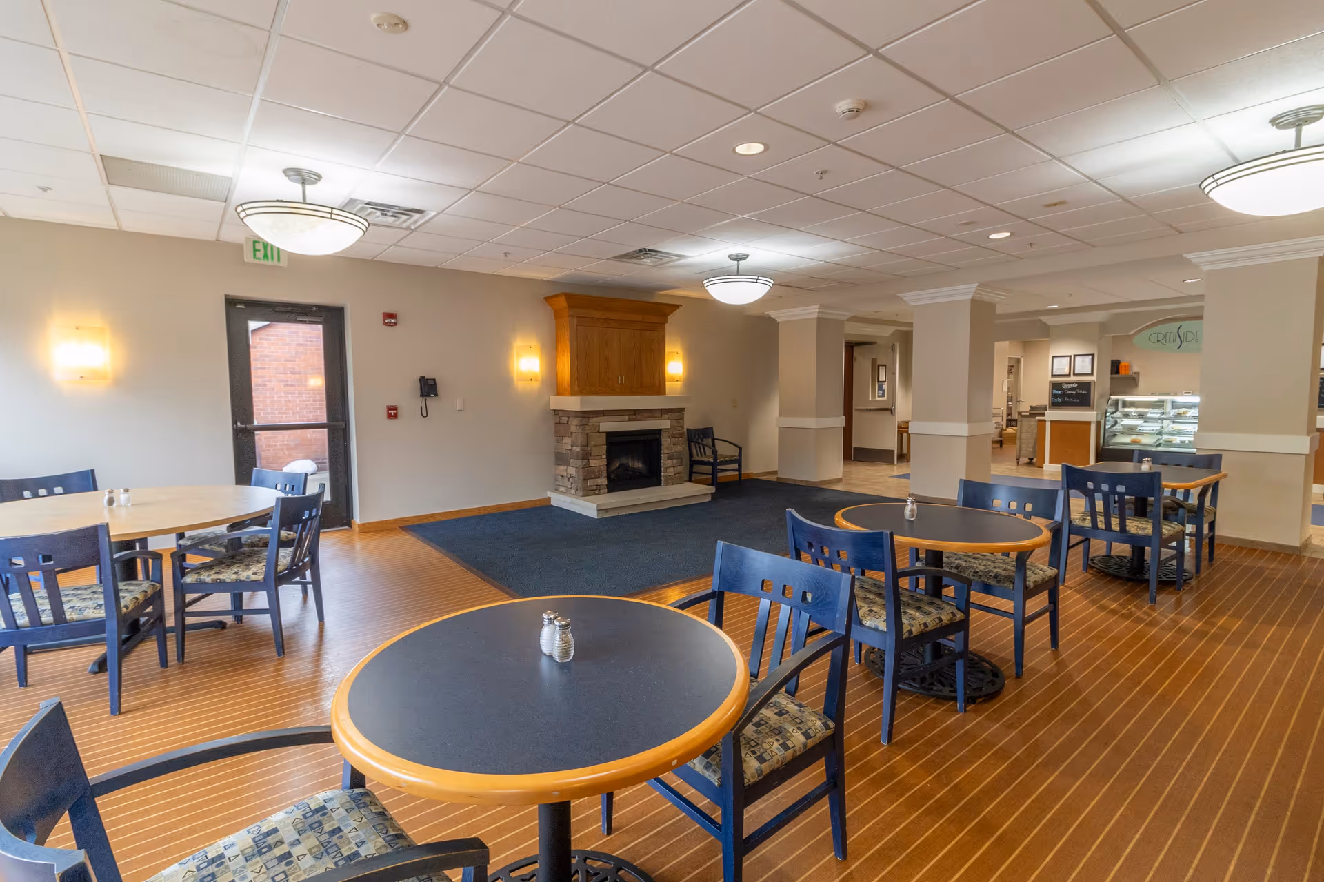 A dining area in a senior living facility featuring several round tables with blue chairs arranged around them. The room has a wooden floor with a carpeted section in front of a stone fireplace with a wooden mantle. Wall sconces and ceiling lights illuminate the space. In the background, there is a counter area with a display case and a sign that reads 'Creekside'.