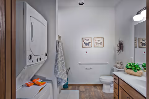 A clean bathroom featuring a stacked washer-dryer on the left, a toilet with grab bar in the center, and a vanity with sink and mirror on the right.