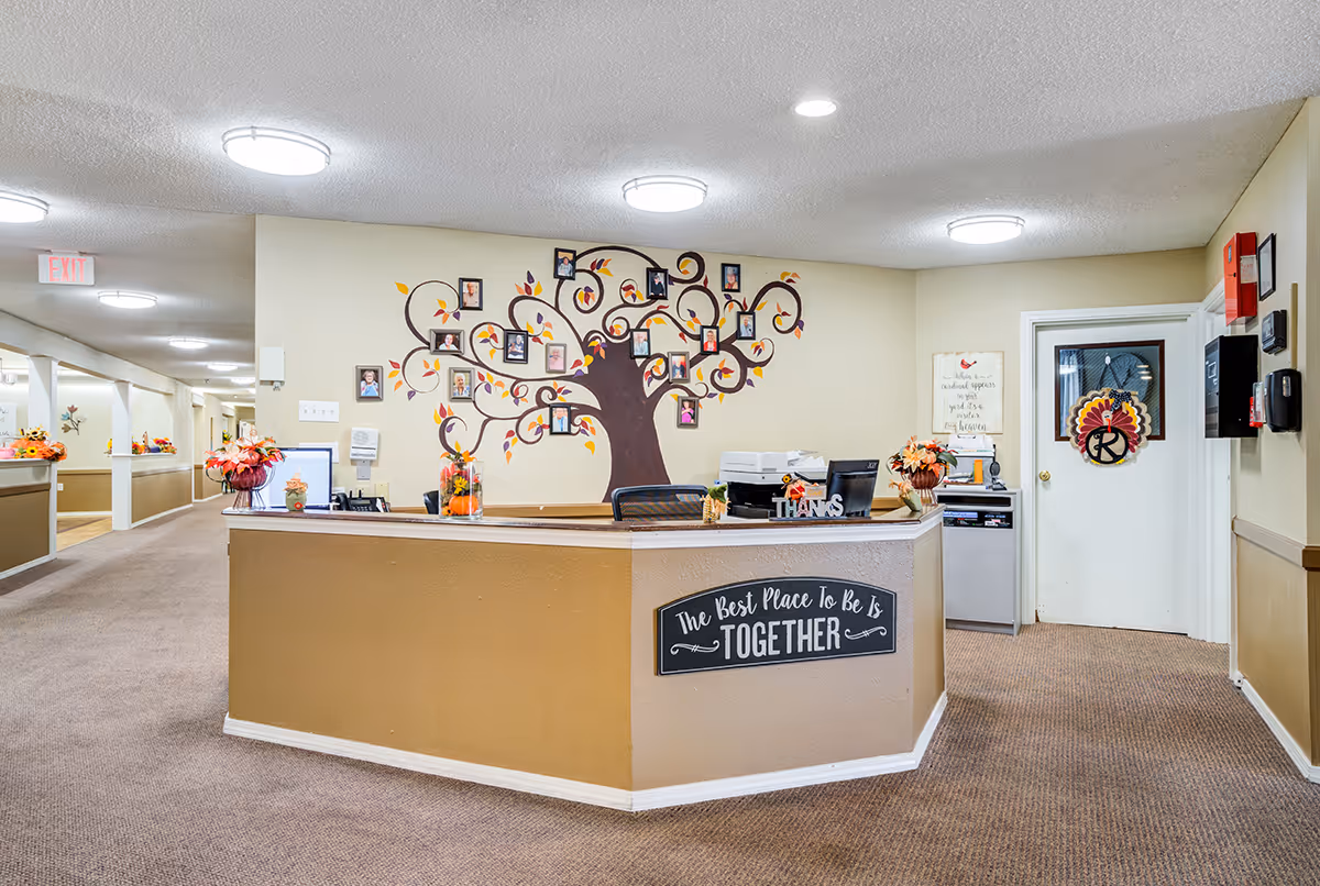 Reception area of Winkler Court facility with a beige and brown front desk decorated with flowers and a sign that reads 'The Best Place To Be Is Together'. Behind the desk is a wall with a large tree mural featuring framed photos. The hallway to the left is well-lit with ceiling lights and decorated with more flowers.