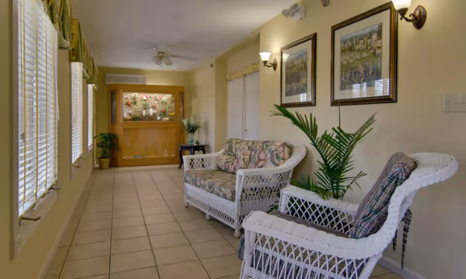 A bright indoor sitting area with white wicker furniture including a loveseat and a chair with floral cushions. The room has tiled flooring, several windows with blinds and valances on the left side, two framed pictures on the right wall, a potted plant, and a ceiling fan. At the far end, there is a wooden display case with decorative items inside.