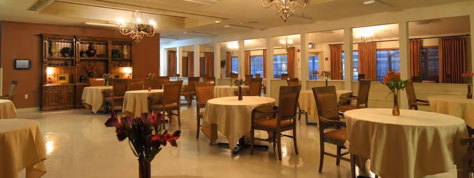 A warmly lit dining room with round tables covered in beige tablecloths, each adorned with a small vase of flowers. Wooden chairs surround the tables, and the room features chandeliers hanging from the ceiling. There is a wooden cabinet with decorative items against one wall and large windows with brown curtains.