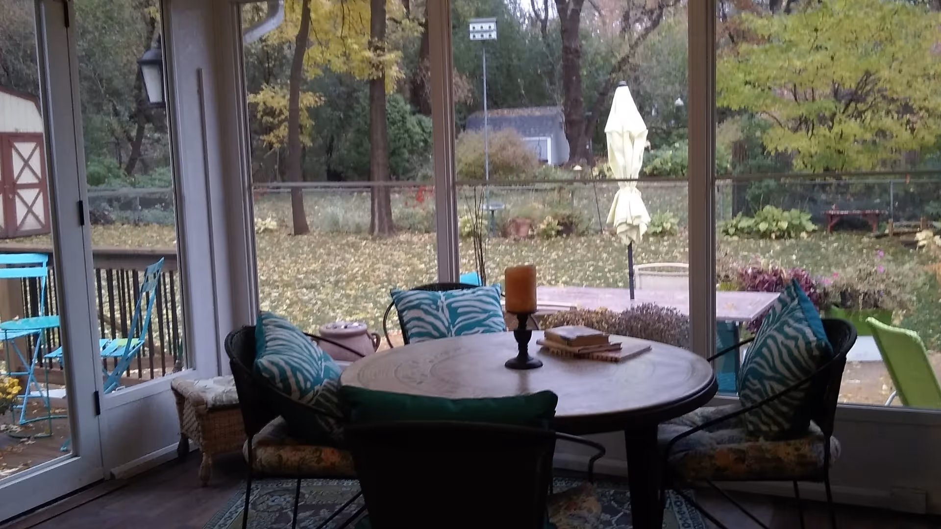 Round wooden dining table with cushioned chairs in a sunroom overlooking a leafy backyard.