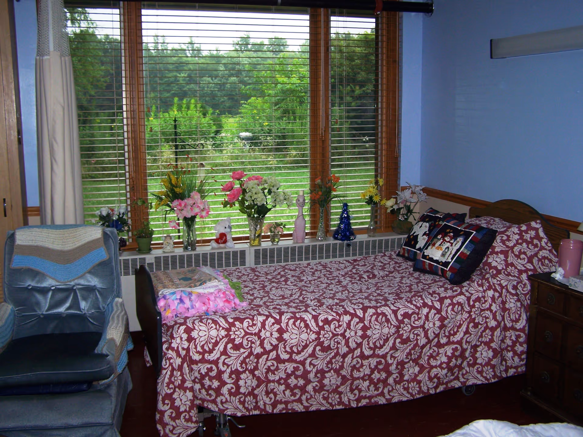 A cozy bedroom with a single bed covered in a red patterned bedspread, a window with blinds and flower arrangements on the windowsill, and a blue armchair.