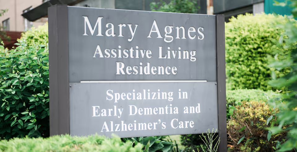 Outdoor sign for "Mary Agnes Assistive Living Residence" stating it specializes in early dementia and Alzheimer's care, surrounded by greenery.
