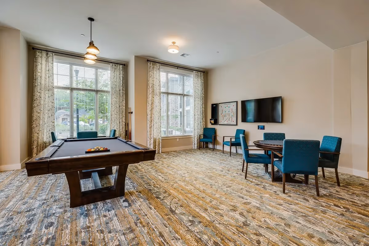 A well-lit recreational room with a pool table in the center, a round table with four blue upholstered chairs, large windows with patterned curtains allowing natural light, a wall-mounted TV, and a framed board game on the wall.