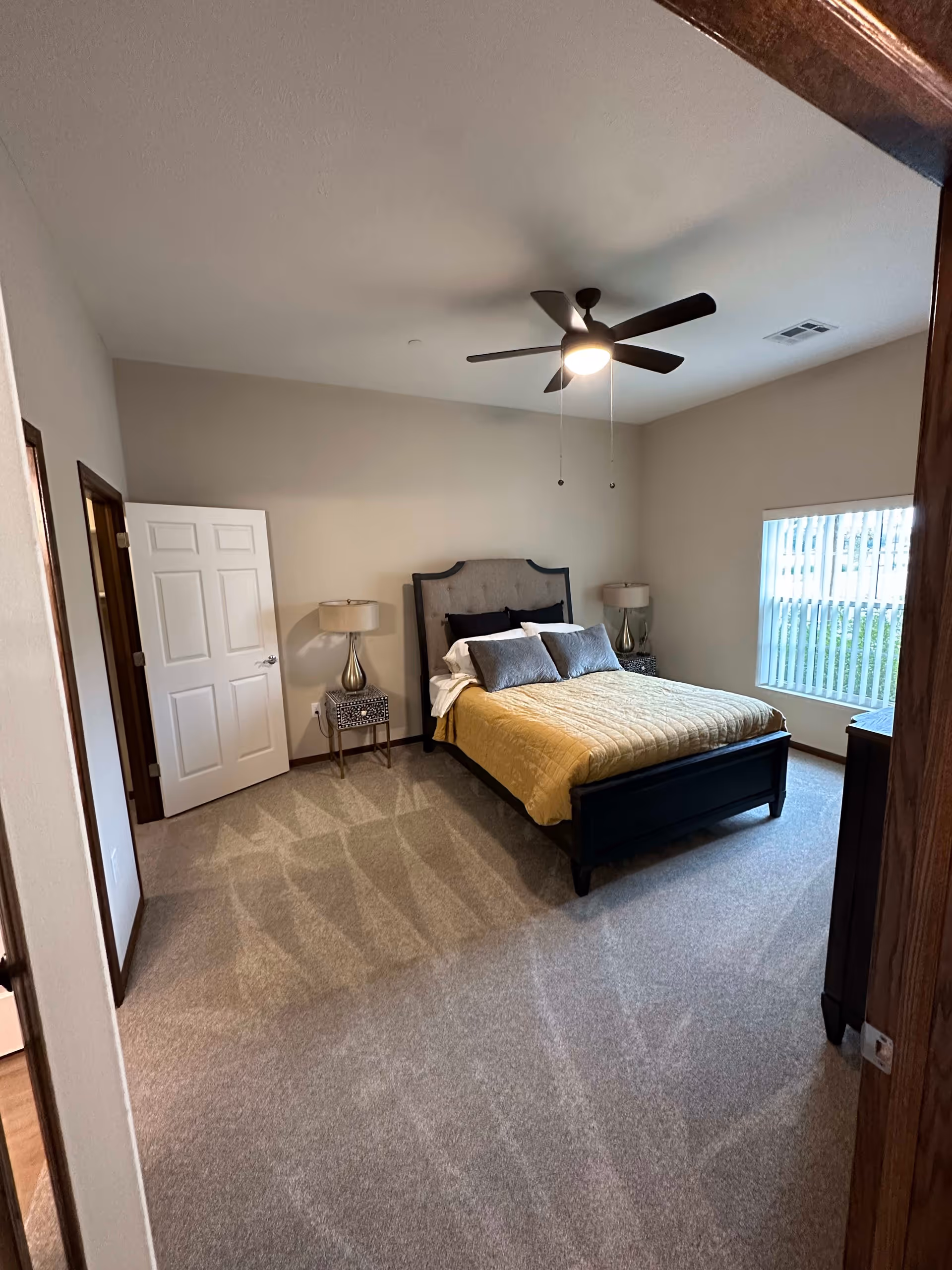 A bedroom with a bed featuring a gray upholstered headboard and yellow bedspread, two bedside tables with lamps, a ceiling fan with light, a window with vertical blinds, and beige carpeted floor.