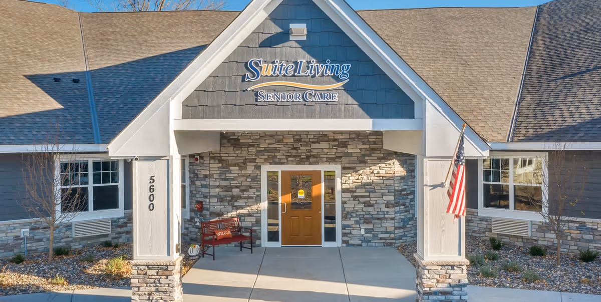 Front entrance of Suite Living Senior Care facility with a peaked roof, stone facade, a wooden door, a red bench with a welcome pillow, and an American flag on the right side.