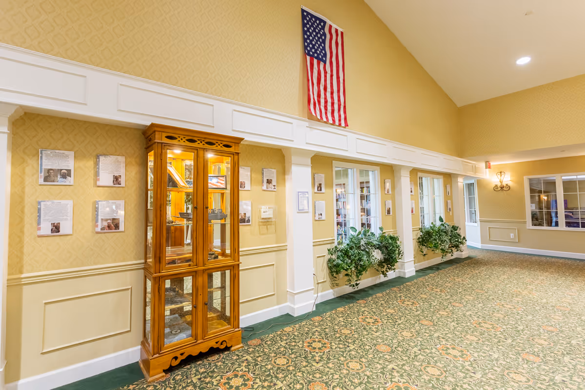 Interior hallway of a senior living facility with patterned green carpet and yellow wallpaper. A wooden display cabinet with glass doors is positioned against the wall, containing various items. Several framed documents and photos are mounted on the wall. Two windows with green plants on the ledges look into another room. An American flag hangs high on the wall above the display cabinet. The ceiling is vaulted with recessed lighting.