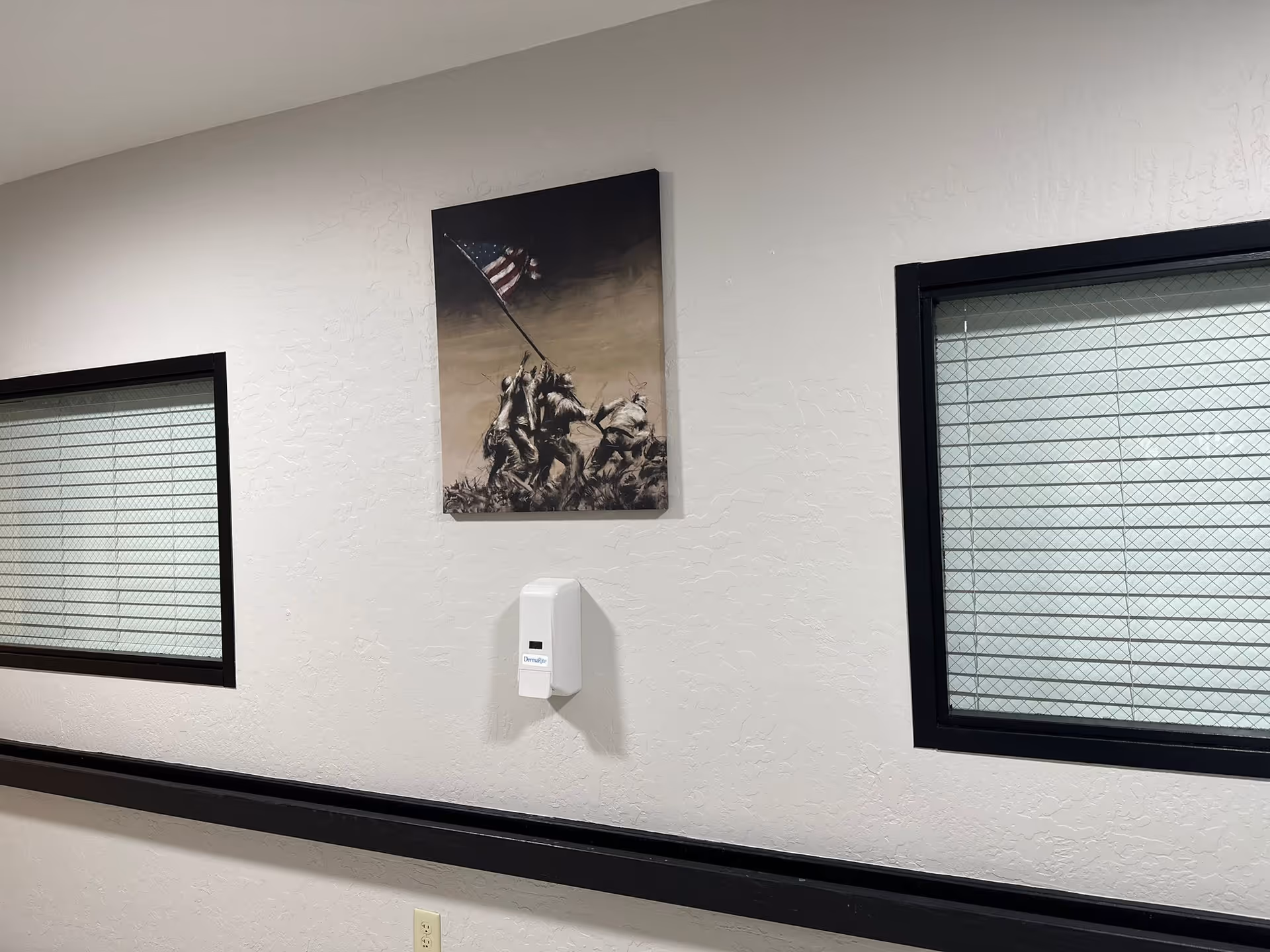 Interior hallway with two windows featuring wire-reinforced glass on either side of a wall. A black handrail runs along the wall below the windows. A wall-mounted hand sanitizer dispenser is centered beneath a framed picture depicting soldiers raising an American flag.