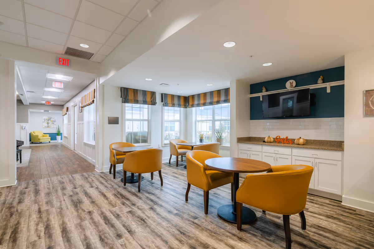 A bright and modern common area with round wooden tables and mustard yellow chairs. Large windows with striped valances let in natural light. A TV is mounted on a teal accent wall above a countertop with white cabinets. The space has wood-look flooring and a hallway with additional seating and artwork is visible in the background.