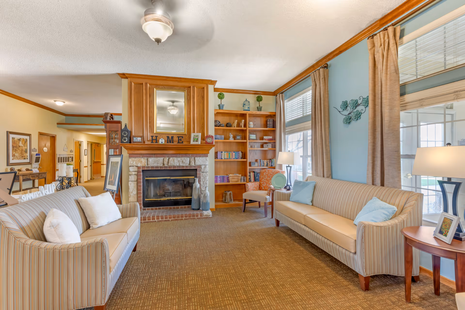 Bright common living room with two striped sofas facing a fireplace and built-in bookshelves, large windows, and lamps.