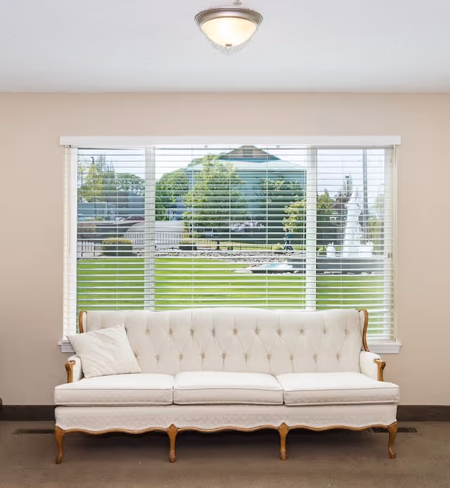 A beige tufted sofa with wooden legs and armrests is placed in front of a large window with white blinds. Outside the window, there is a green lawn, trees, and a water fountain. The room has beige walls and a ceiling light fixture.