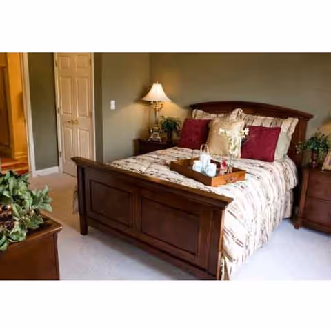 A cozy bedroom featuring a wooden bed with a patterned bedspread and red accent pillows. A wooden nightstand with a lamp and decorative items is beside the bed. There is a door slightly ajar leading to another room, and a green plant is visible on a piece of furniture in the foreground.
