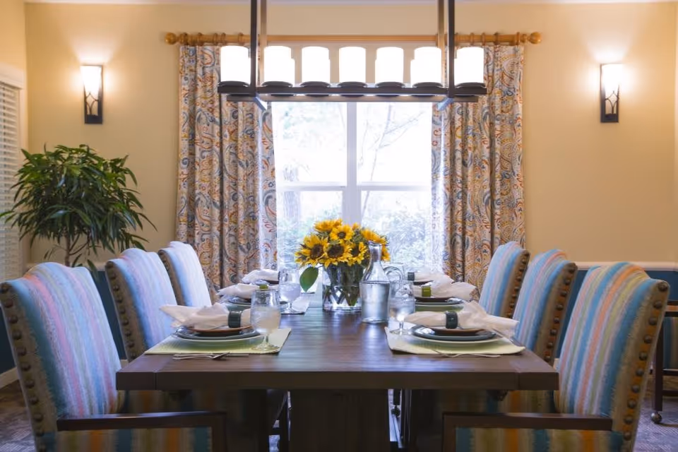 A dining room with a wooden table set for six people. The table has plates, glasses, napkins, and a centerpiece of sunflowers in a vase. The chairs around the table have colorful striped upholstery. Behind the table is a window with patterned curtains, and there are two wall sconces providing light. A green plant is visible in the corner of the room.