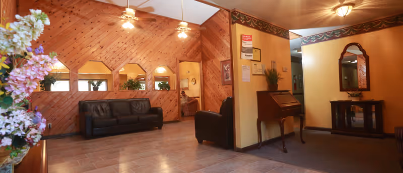 Wood-paneled senior living facility lobby with leather sofas, a small writing desk, mirror, and floral arrangements.