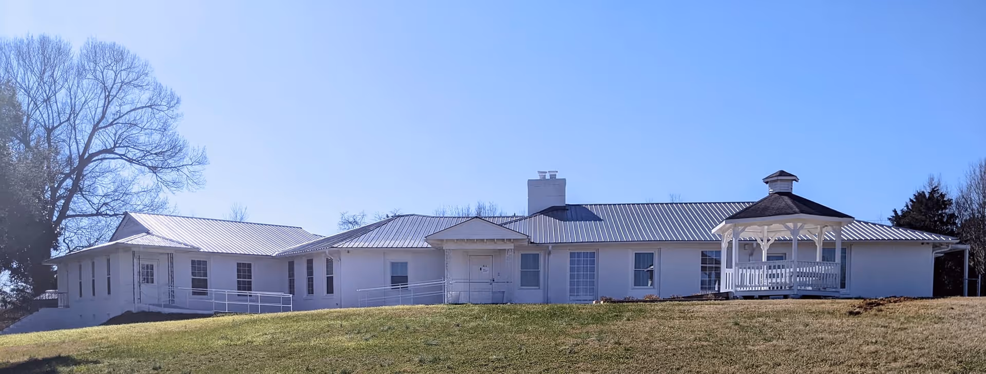 Exterior view of a single-story white building with a metal roof, a central chimney, and a white gazebo on the right side. The building is situated on a grassy hill with leafless trees in the background under a clear blue sky.