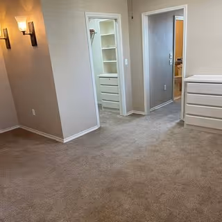 An empty room with beige carpet and light beige walls featuring two wall-mounted light fixtures. There are two doorways visible, one leading to a closet with built-in shelves and drawers, and the other leading to another room with a glimpse of a kitchen area. A white dresser is positioned against the right wall.