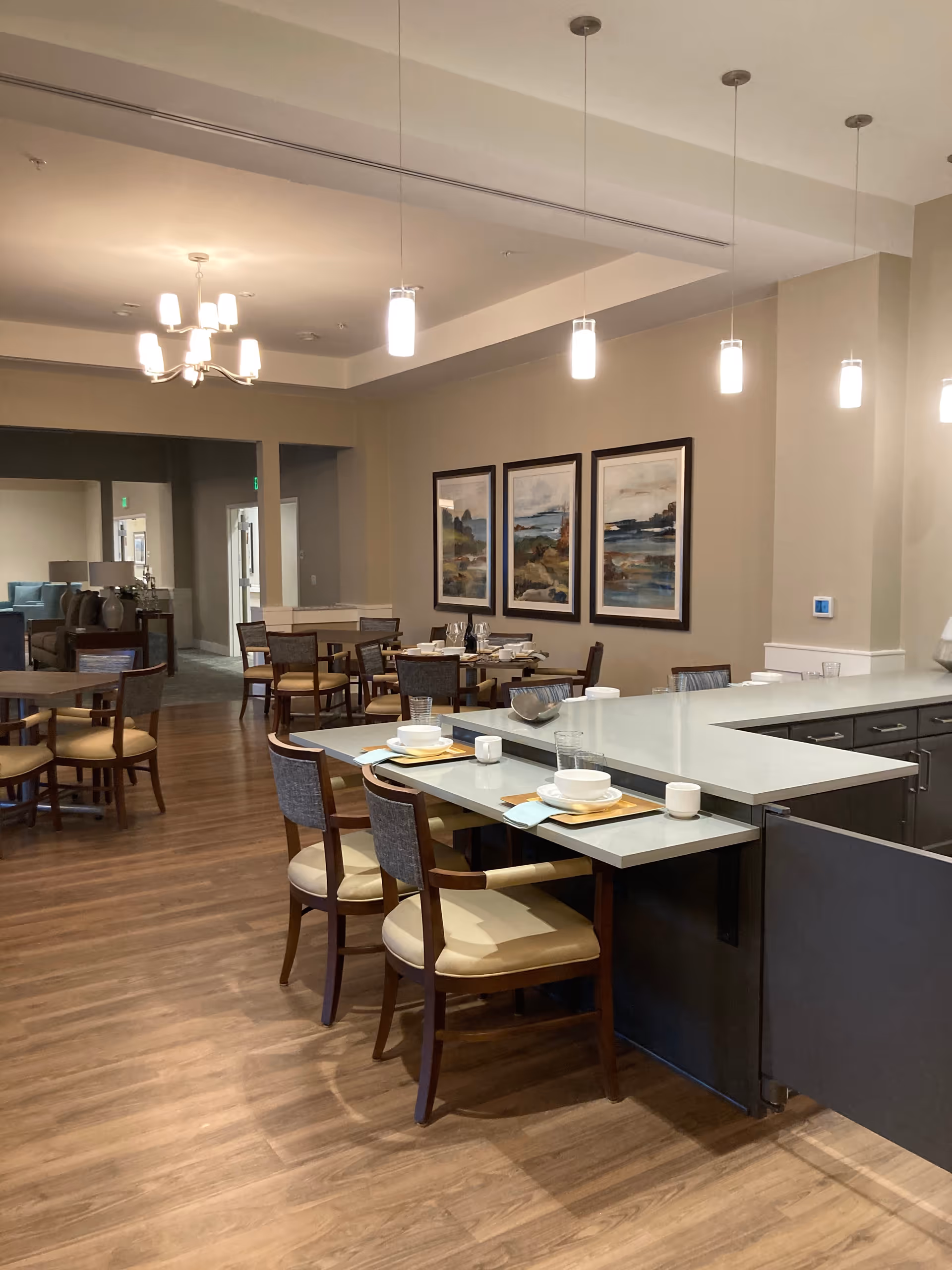 A dining area in an assisted living facility with several tables and chairs arranged neatly. The tables are set with plates, bowls, cups, and napkins. The room features wooden flooring, modern pendant lights hanging from the ceiling, and three framed landscape paintings on the wall. In the background, there is a glimpse of a lounge area with sofas.