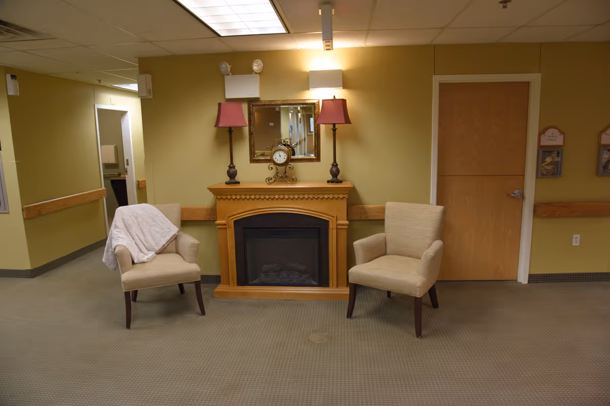 A cozy seating area in a hallway with two beige armchairs placed on either side of a wooden electric fireplace. One chair has a white throw blanket draped over it. Above the fireplace is a decorative clock and two lamps with red shades, and a mirror reflecting the hallway. The walls are painted light green, and there is a wooden door and hallway visible in the background.