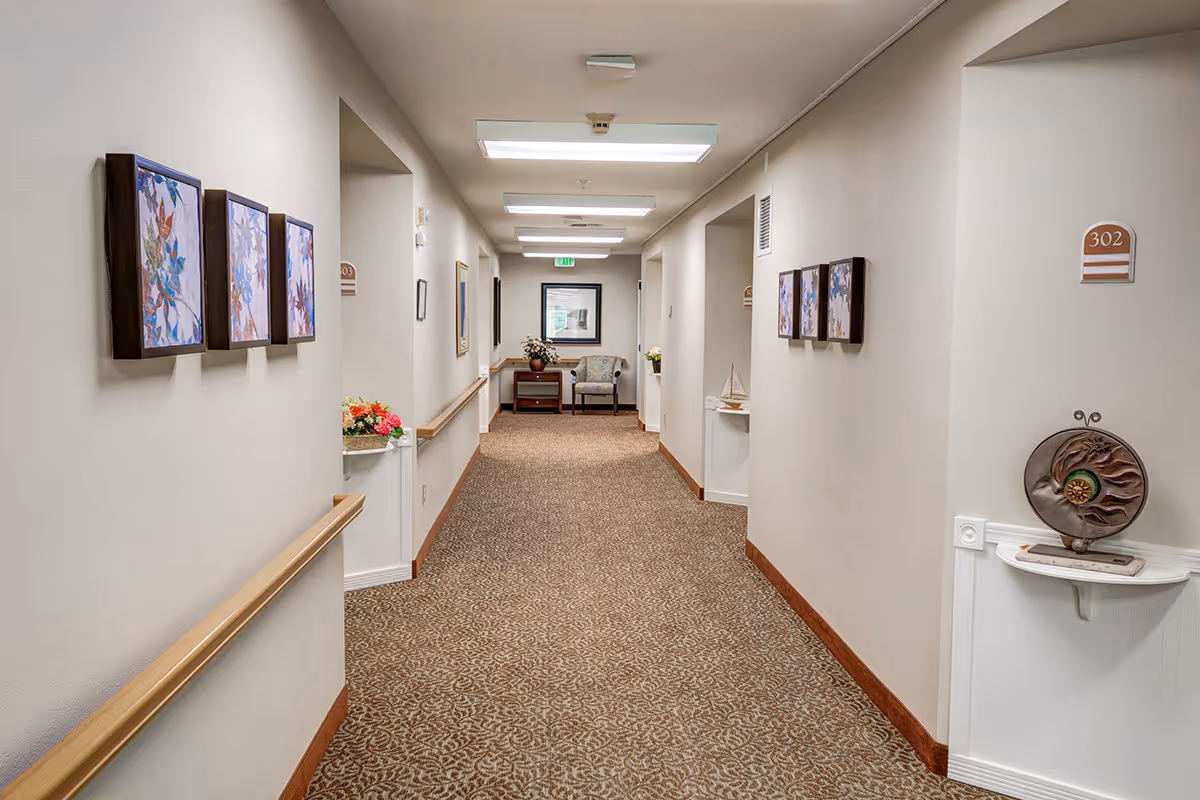 A well-lit hallway in a senior living facility with beige walls and patterned carpet. The hallway features handrails on both sides, framed floral artwork on the walls, and small decorative shelves with flowers and a sculpture. At the end of the hallway, there is a chair and a table with a flower arrangement beneath a framed picture.