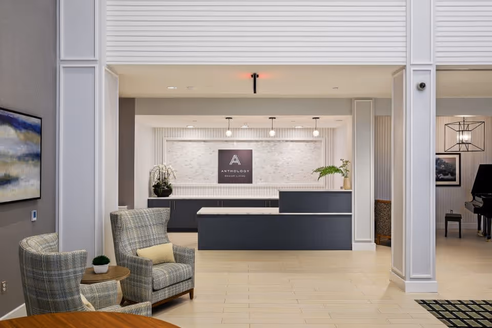 A modern reception area with a dark blue front desk and a white marble countertop. Behind the desk is a wall with a sign that reads 'Anthology Senior Living' and decorative lighting hanging from the ceiling. To the left, there are two plaid upholstered armchairs with a small round wooden table between them, holding a small potted plant. The floor is light-colored tile, and the walls are painted in neutral tones. On the right side, part of a piano and a framed picture are visible.