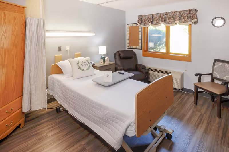 A clean senior bedroom featuring an adjustable hospital-style bed with white linens, a bedside table and lamp, a recliner, and a window.
