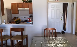 Dining area with glass table and wooden chairs; kitchen and front door visible in background.