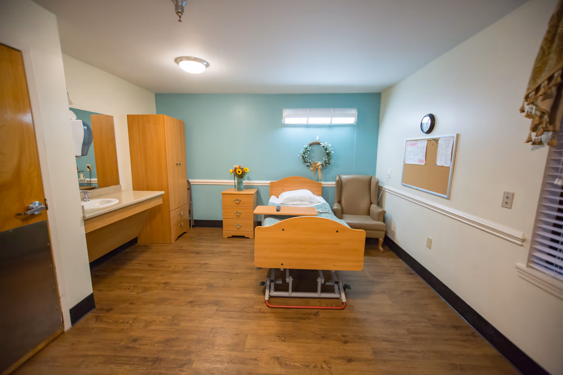 A simple healthcare center bedroom with a single adjustable bed, a wooden nightstand with a vase of flowers, a comfortable armchair, a wall clock, a bulletin board, and a sink with a mirror. The walls are painted light blue and white, and the floor has wood-like flooring.