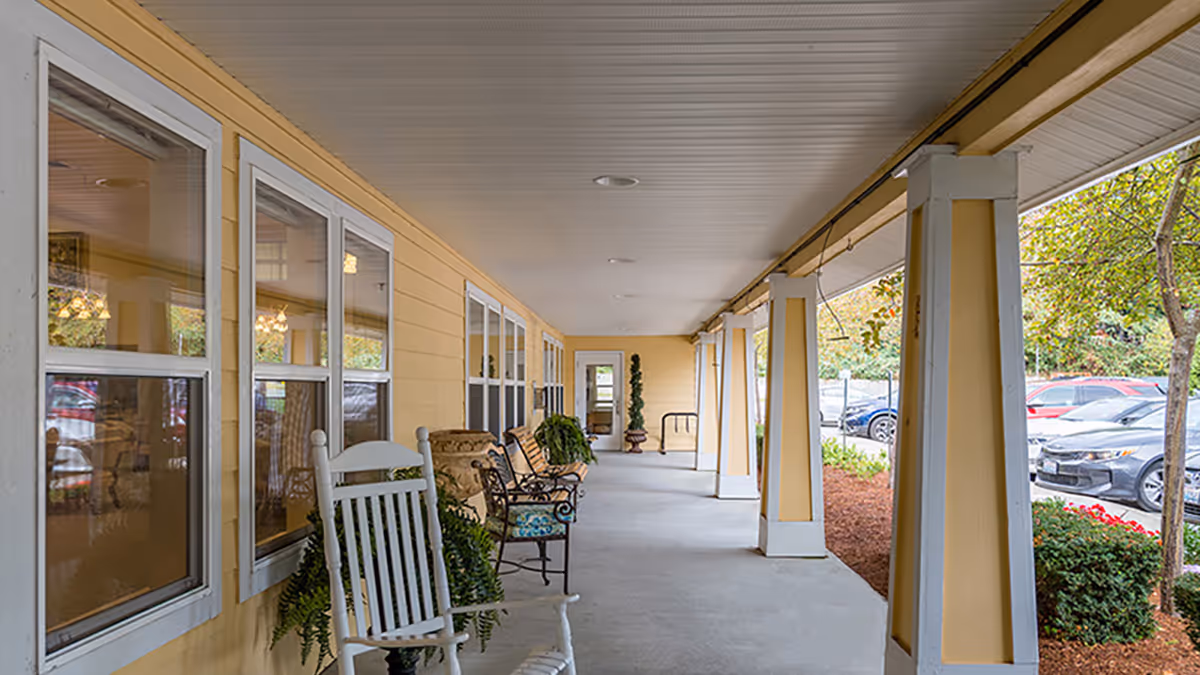 Covered yellow-painted front porch with rocking chairs, benches, and columns overlooking a parking lot.