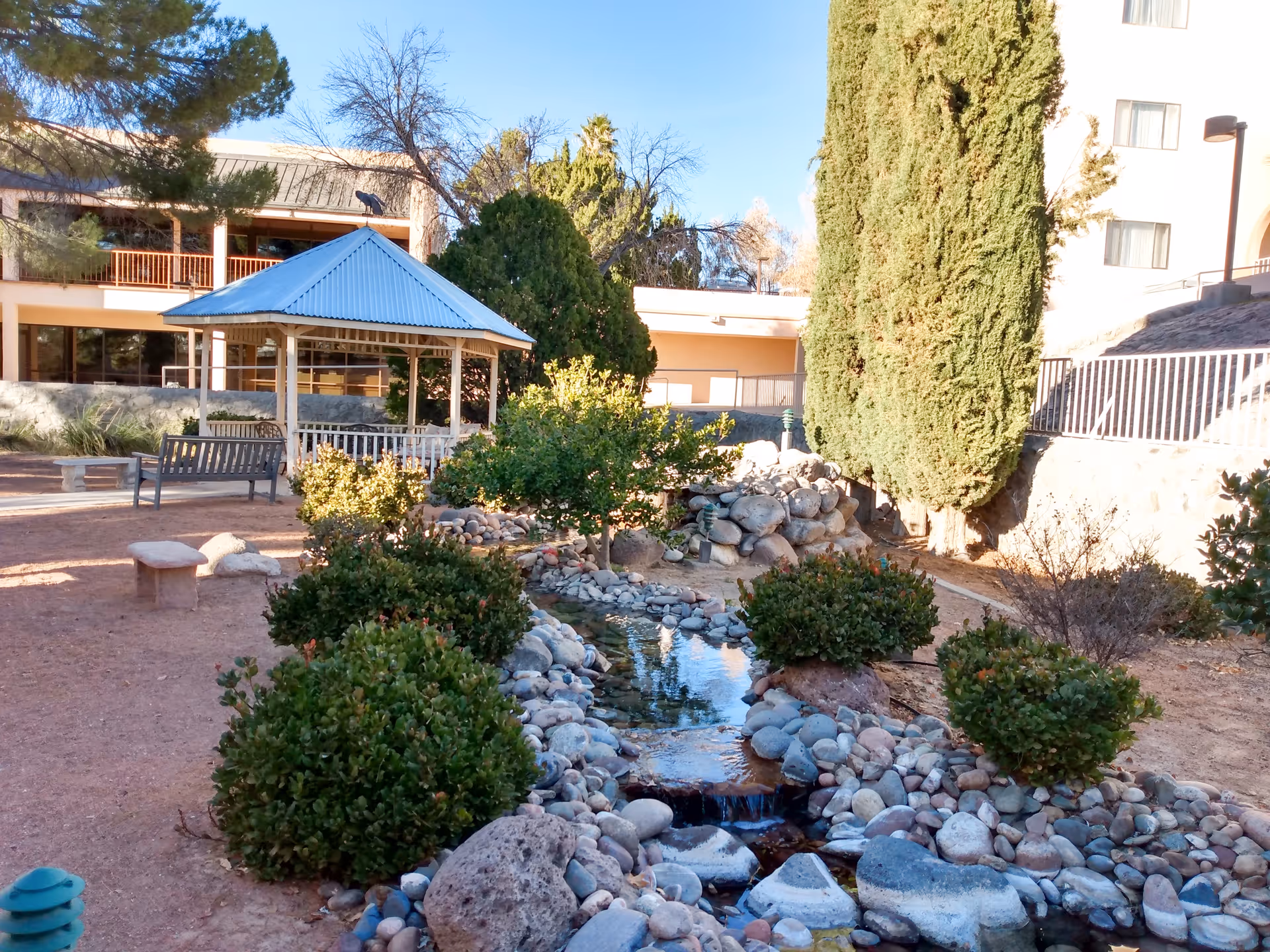 A landscaped outdoor courtyard with a rock-lined stream, shrubs, benches, and a gazebo next to the facility building.