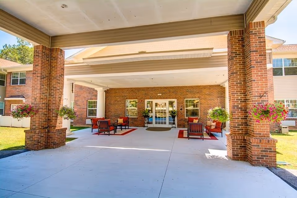 Covered outdoor entrance area of a senior living facility with brick pillars, hanging flower baskets, and seating arrangements with red cushions on rugs near the entrance doors.