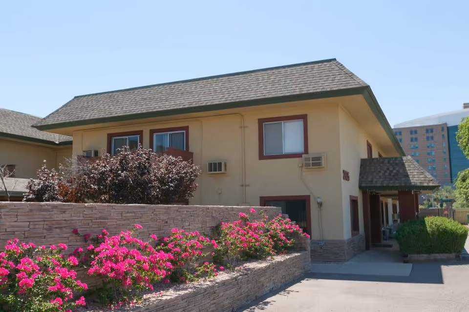 Exterior view of a two-story senior living facility building with beige walls and a shingled roof. There are windows with air conditioning units and a stone retaining wall in front with vibrant pink flowers and shrubs. The sky is clear and blue.